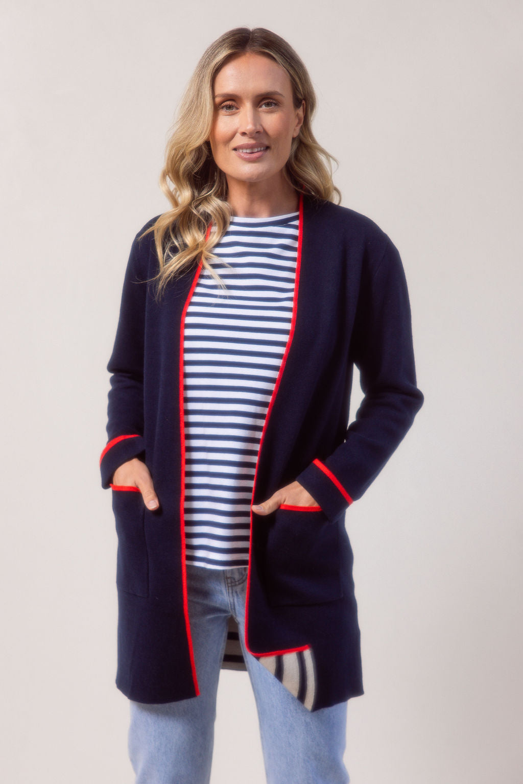 Woman wearing a See Saw navy cardigan with red trim, striped shirt, jeans, and white sneakers on a plain background