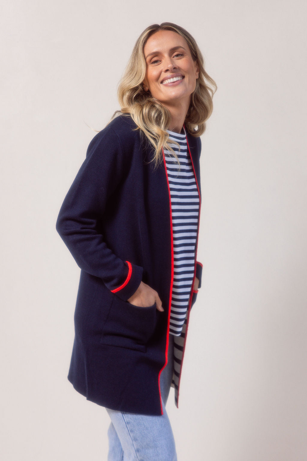 Woman wearing a See Saw navy cardigan with red trim, striped shirt, jeans, and white sneakers on a plain background