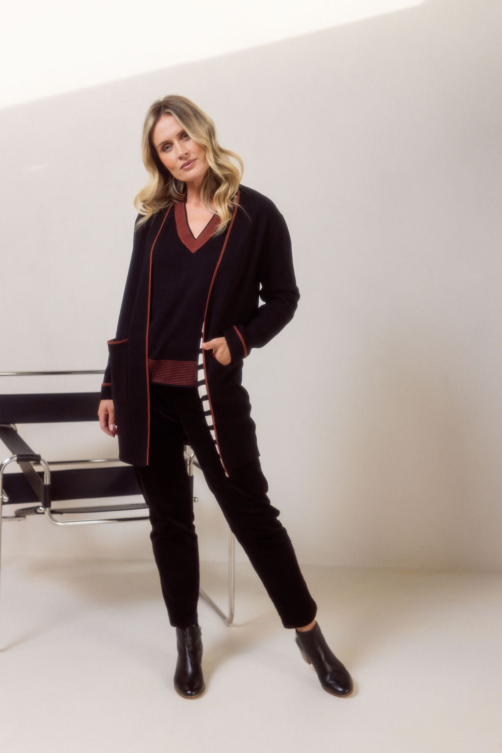 Woman wearing a See Saw black cardigan with copper trim, black sweater and pants and black boots on a plain background