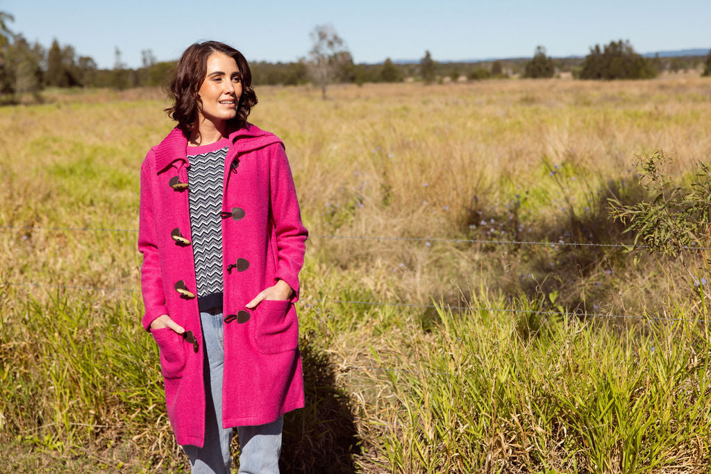 Woman wearing a pink See Saw Duffle coat in boiled wool standing in a field with trees and sky in the background