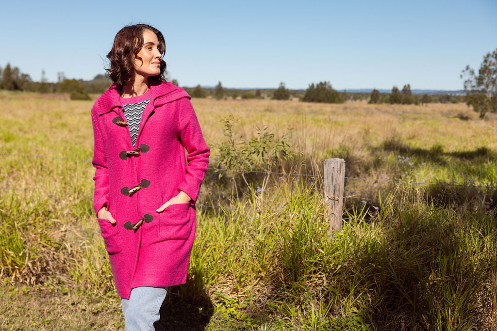Woman wearing a pink See Saw Duffle coat in boiled wool standing in a field with trees and sky in the background