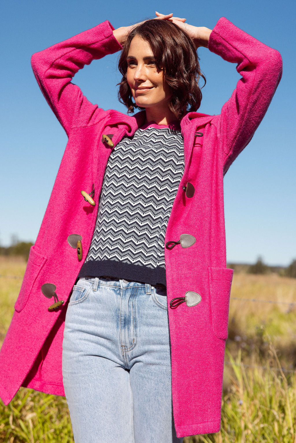 Woman wearing a pink See Saw Duffle coat in boiled wool standing in a field with trees and sky in the background