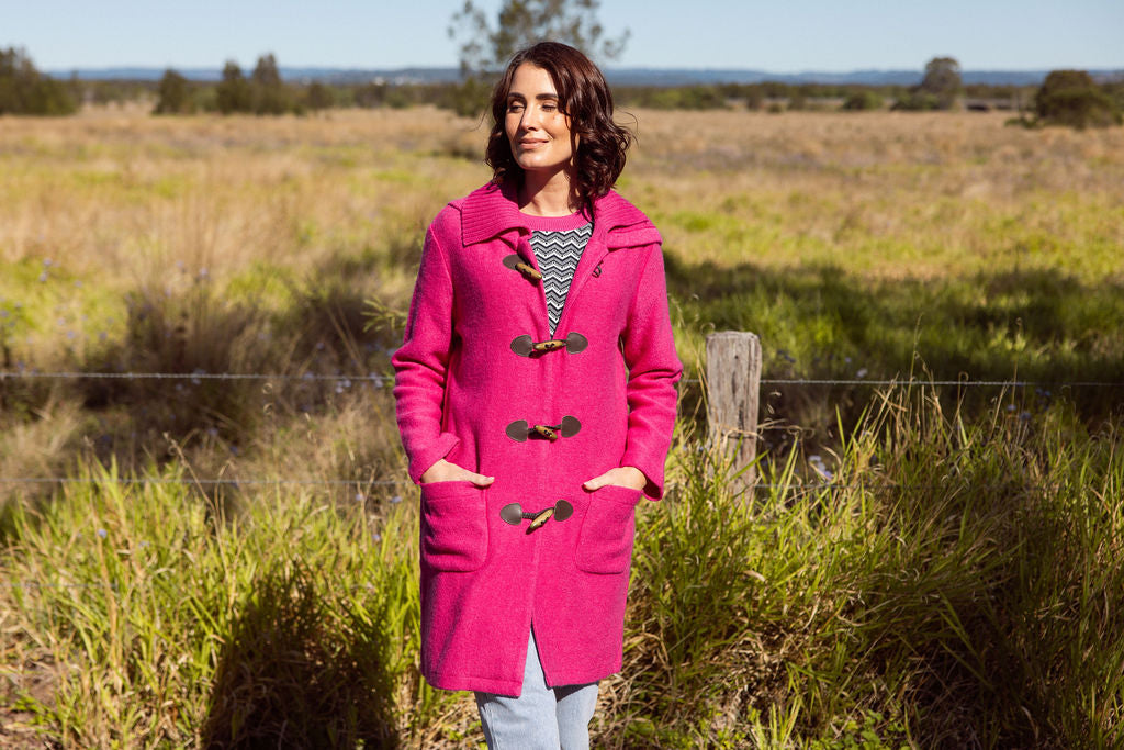 Woman wearing a pink See Saw Duffle coat in boiled wool standing in a field with trees and sky in the background