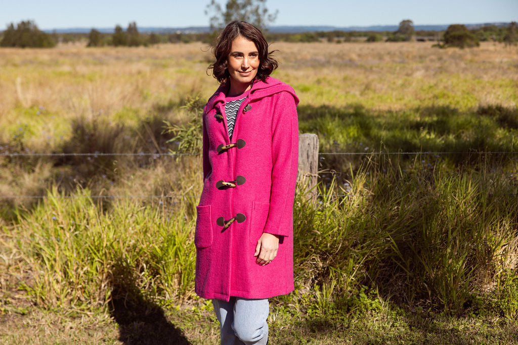 Woman wearing a pink See Saw Duffle coat in boiled wool standing in a field with trees and sky in the background