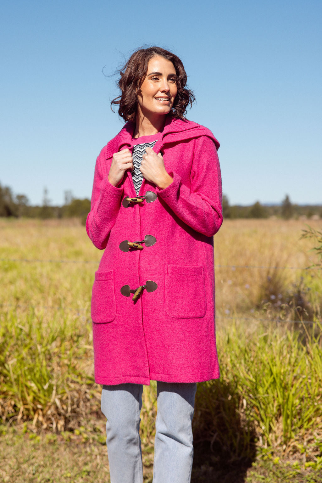 Woman wearing a pink See Saw Duffle coat in boiled wool standing in a field with trees and sky in the background