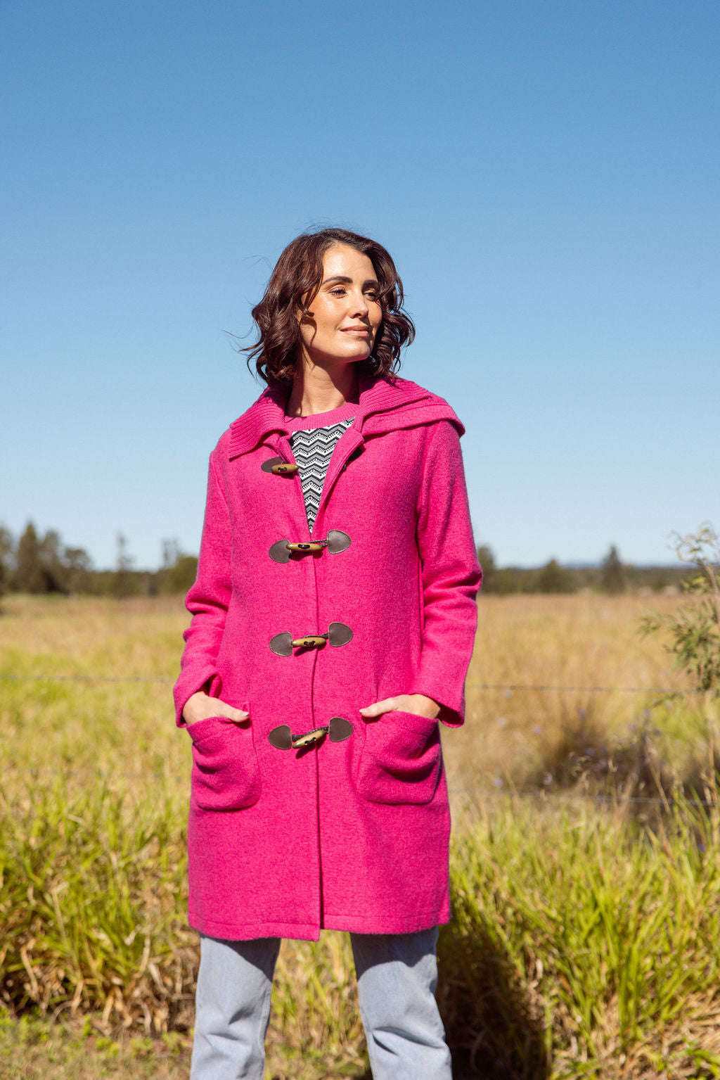 Woman wearing a pink See Saw Duffle coat in boiled wool standing in a field with trees and sky in the background
