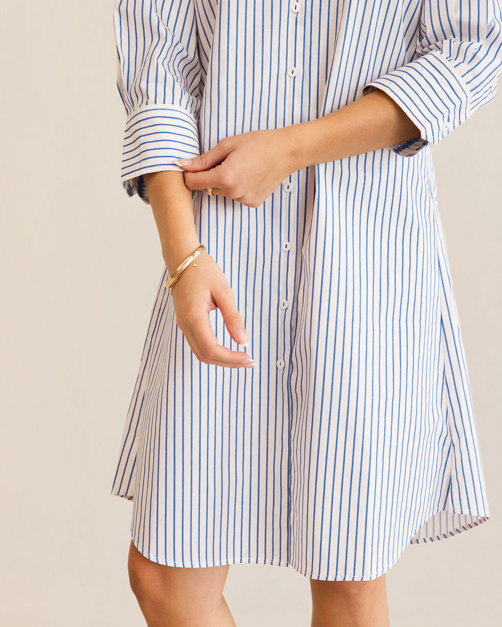 A woman modeling a blue and white Kaleido cotton vertically striped button-down shirt dress with three-quarter sleeves, standing against a plain off-white studio background.