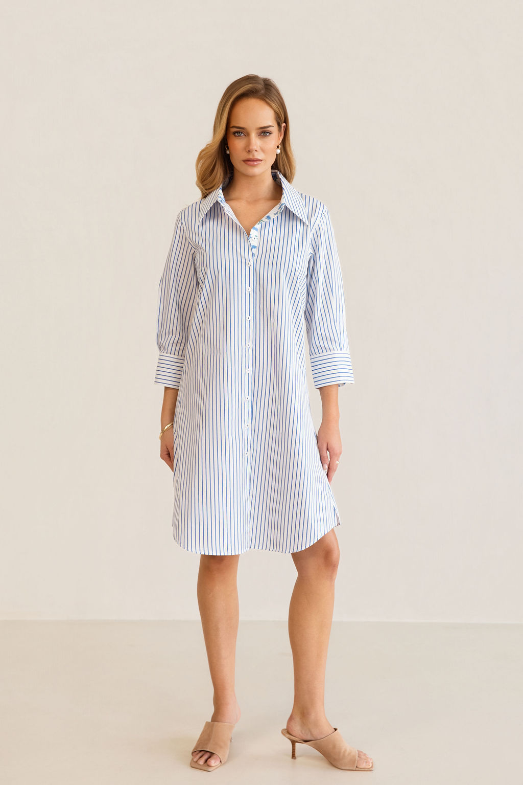 A woman modeling a blue and white Kaleido cotton vertically striped button-down shirt dress with three-quarter sleeves, standing against a plain off-white studio background.