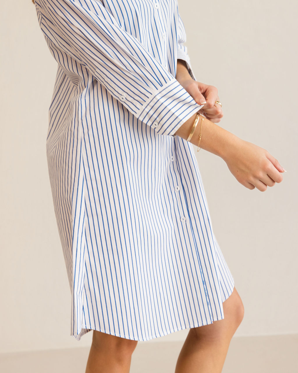 A woman modeling a blue and white Kaleido cotton vertically striped button-down shirt dress with three-quarter sleeves, standing against a plain off-white studio background.