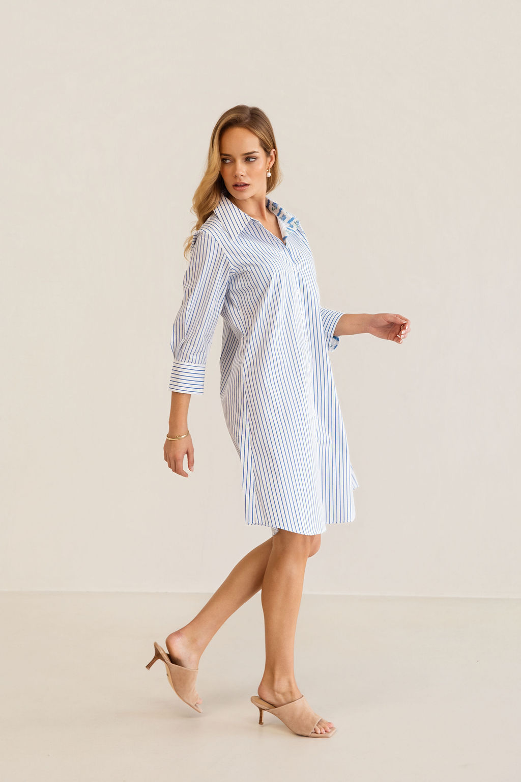 A woman modeling a blue and white Kaleido cotton vertically striped button-down shirt dress with three-quarter sleeves, standing against a plain off-white studio background.