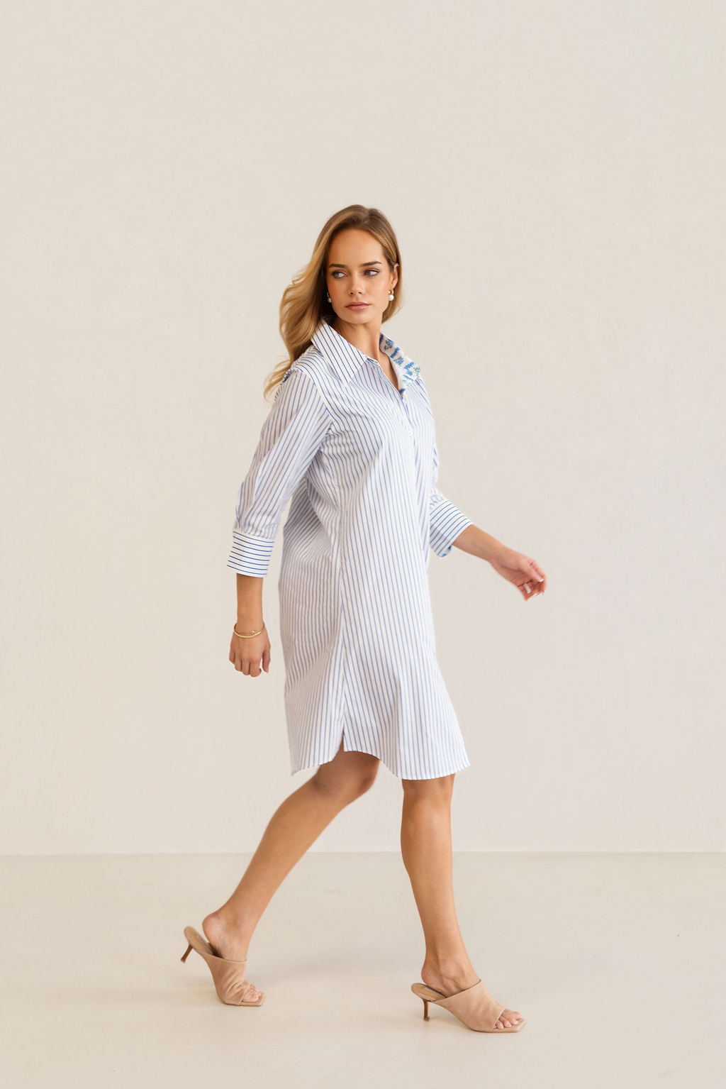 A woman modeling a blue and white Kaleido cotton vertically striped button-down shirt dress with three-quarter sleeves, standing against a plain off-white studio background.