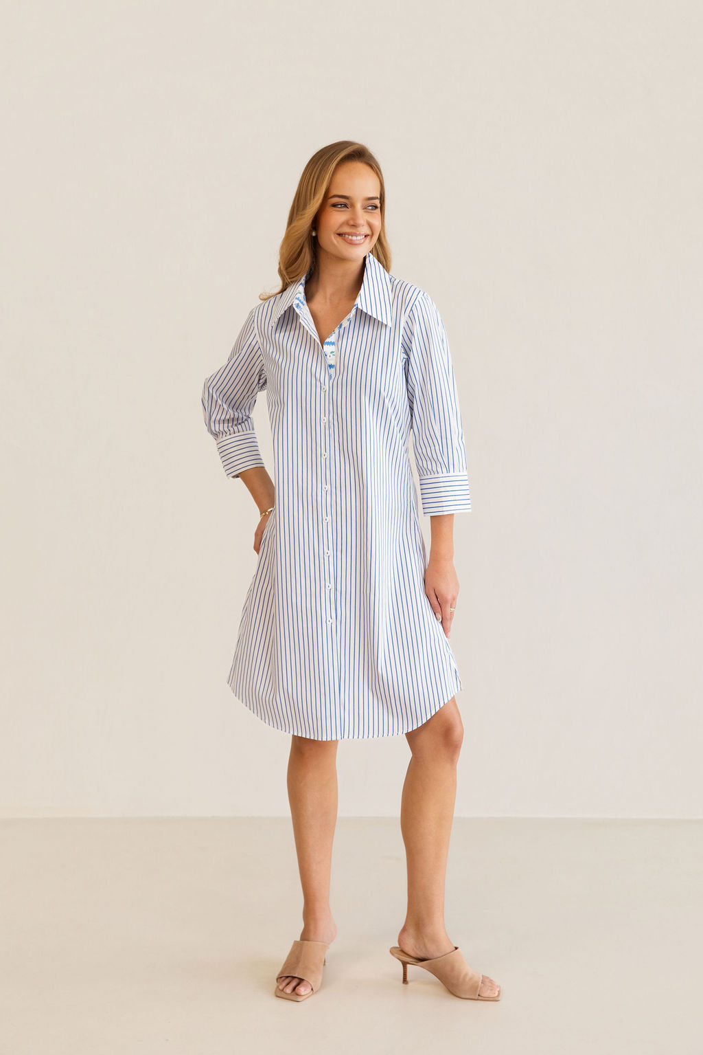 A woman modeling a blue and white Kaleido cotton vertically striped button-down shirt dress with three-quarter sleeves, standing against a plain off-white studio background.