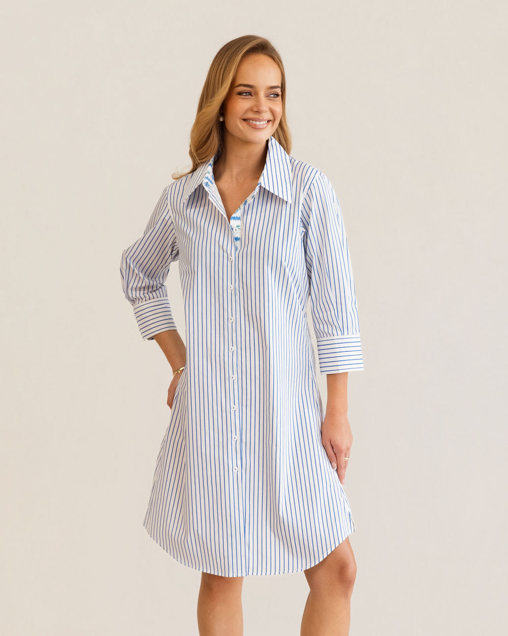 A woman modeling a blue and white Kaleido cotton vertically striped button-down shirt dress with three-quarter sleeves, standing against a plain off-white studio background.