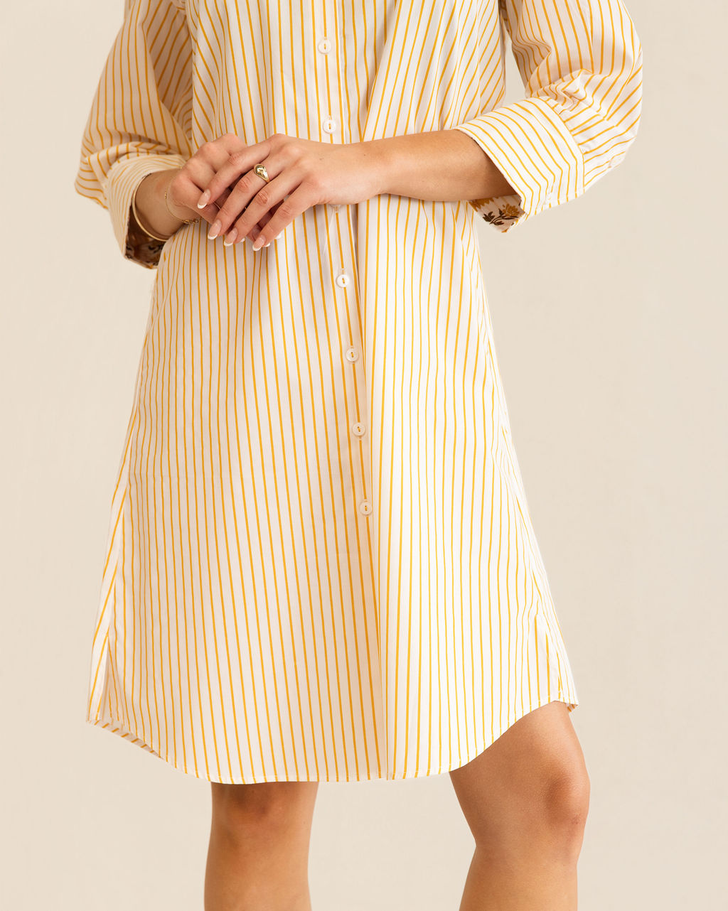 A woman modeling a yellow and white Kaleido cotton vertically striped button-down shirt dress with three-quarter sleeves, standing against a plain off-white studio background.