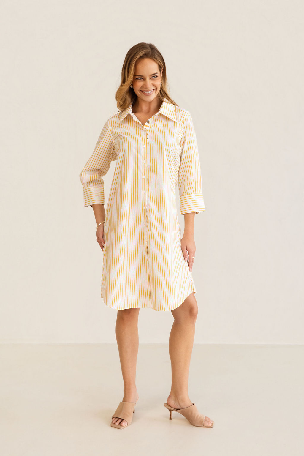 A woman modeling a yellow and white Kaleido cotton vertically striped button-down shirt dress with three-quarter sleeves, standing against a plain off-white studio background.