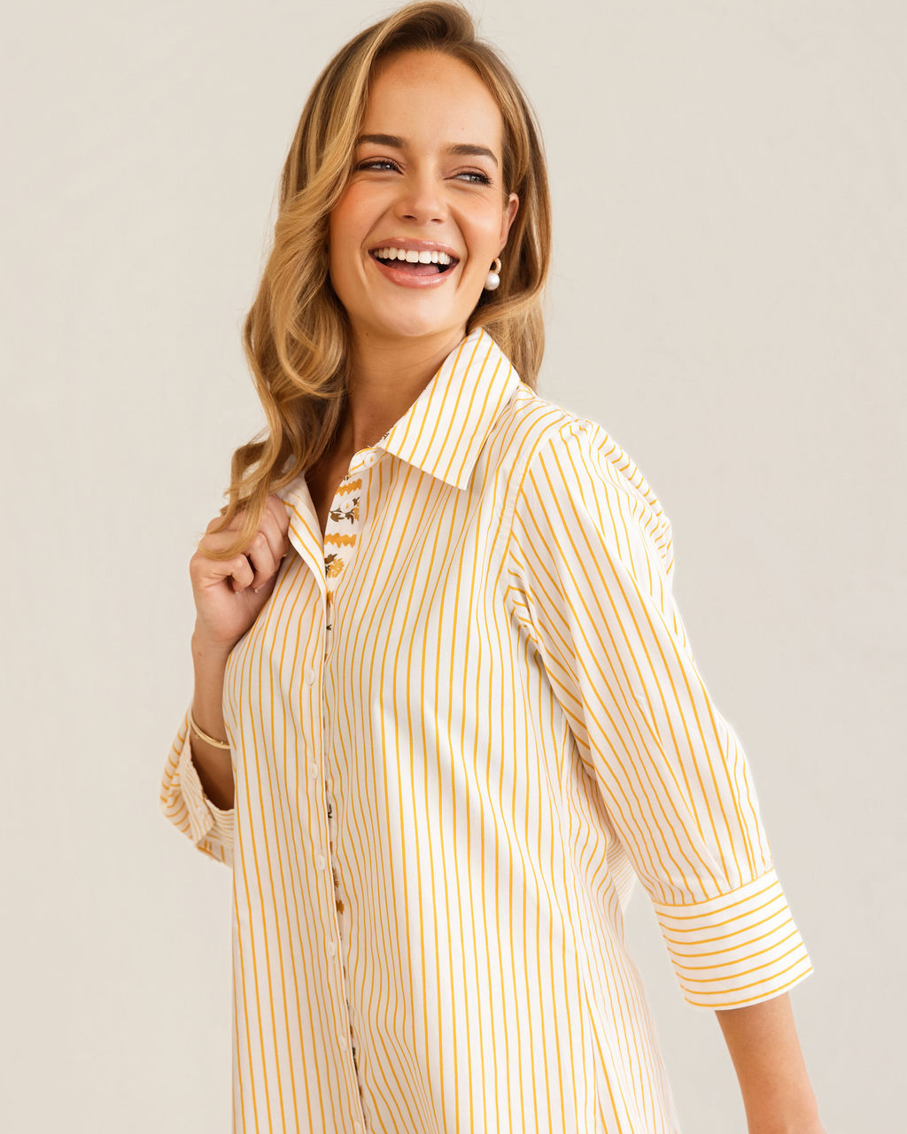 A woman modeling a yellow and white Kaleido cotton vertically striped button-down shirt dress with three-quarter sleeves, standing against a plain off-white studio background.