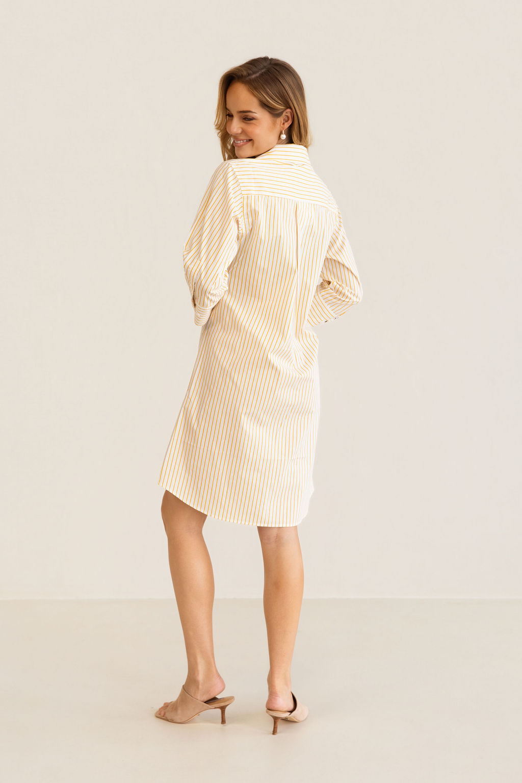 A woman modeling a yellow and white Kaleido cotton vertically striped button-down shirt dress with three-quarter sleeves, standing against a plain off-white studio background.