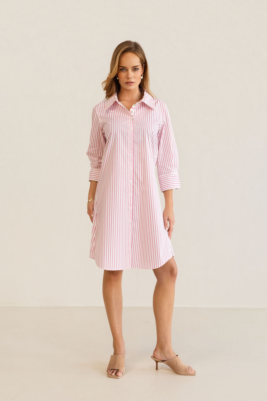 A woman modeling a pink and white Kaleido cotton vertically striped button-down shirt dress with three-quarter sleeves, standing against a plain off-white studio background.