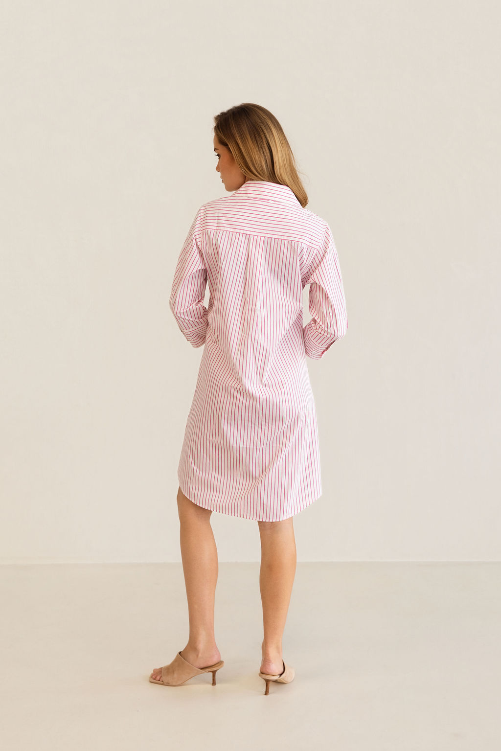 A woman modeling a pink and white Kaleido cotton vertically striped button-down shirt dress with three-quarter sleeves, standing against a plain off-white studio background.