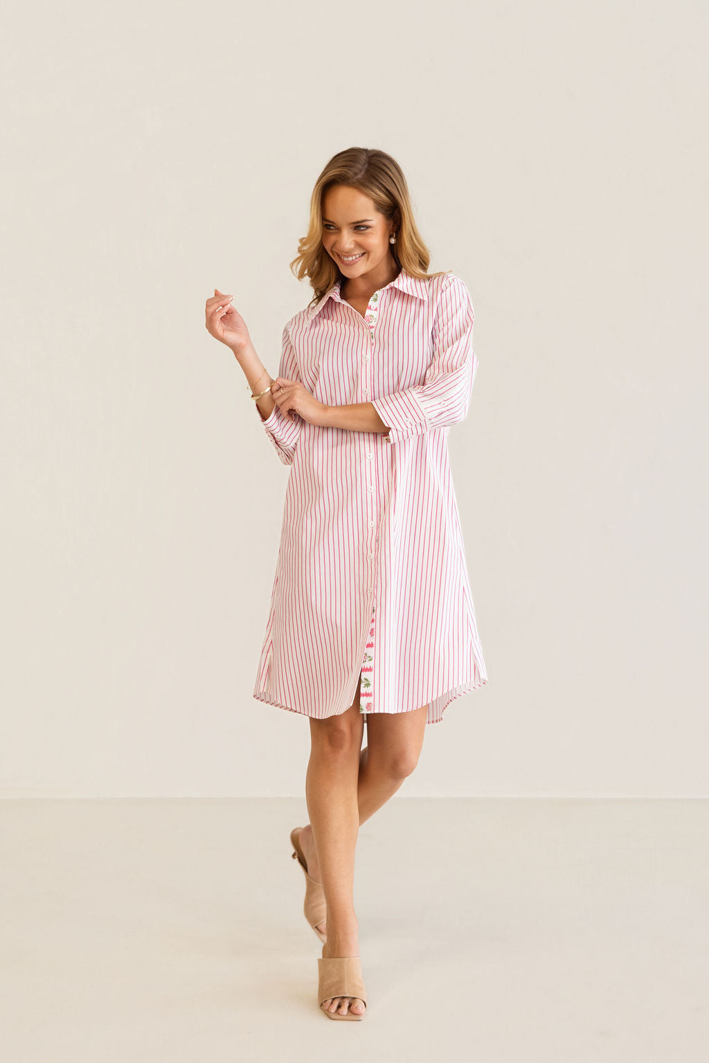 A woman modeling a pink and white Kaleido cotton vertically striped button-down shirt dress with three-quarter sleeves, standing against a plain off-white studio background.