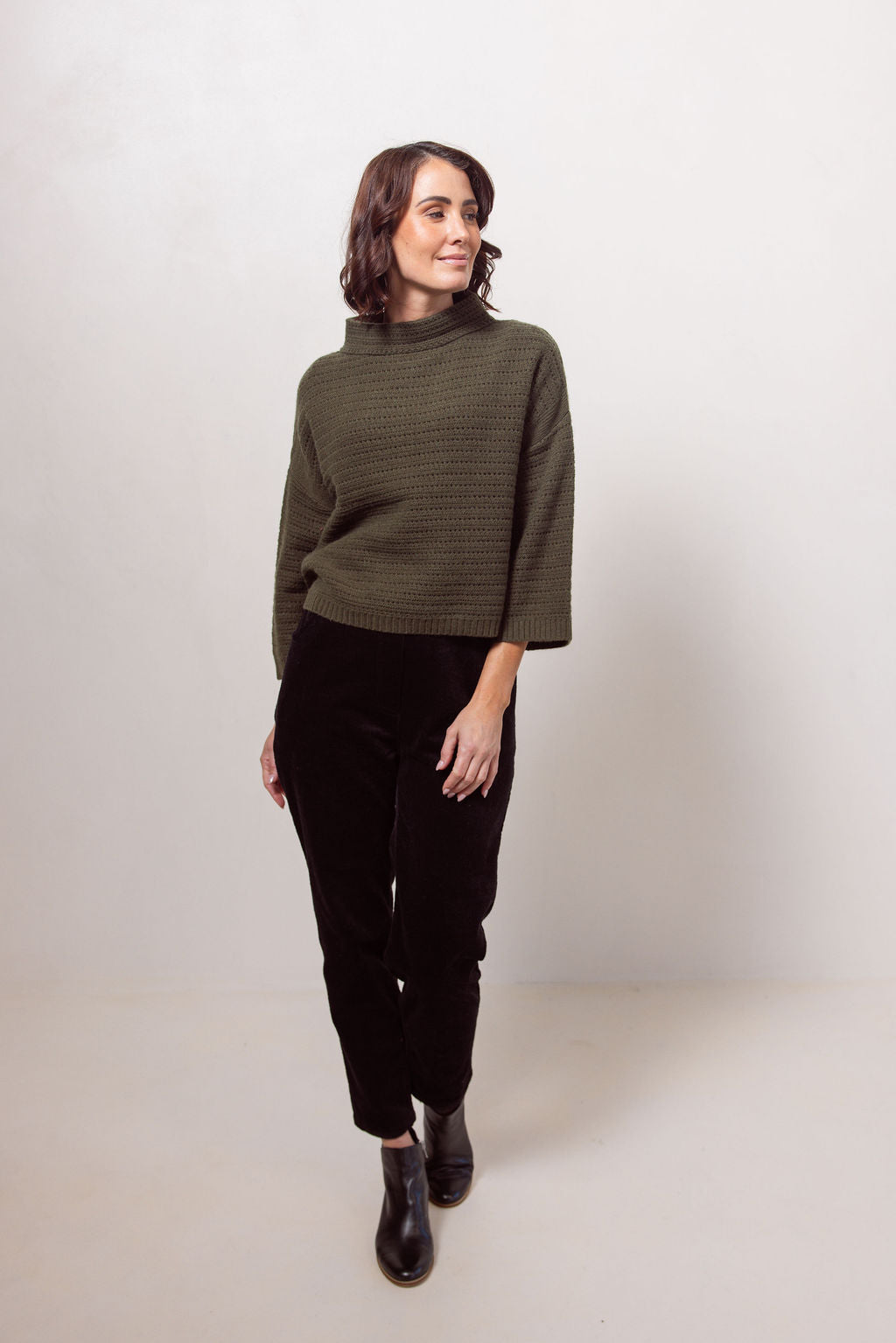 Woman wearing a See Saw Khaki pointelle sweater and black pants standing against a white background.