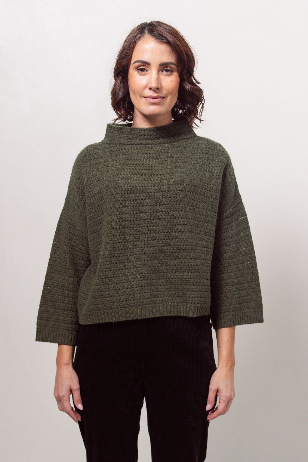 Woman wearing a See Saw Khaki pointelle sweater and black pants standing against a white background.