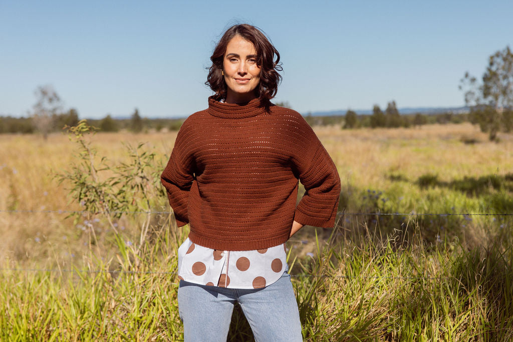 Woman wearing a See Saw copper pointelle sweater and polka dot shirt standing in a field with a clear blue sky.