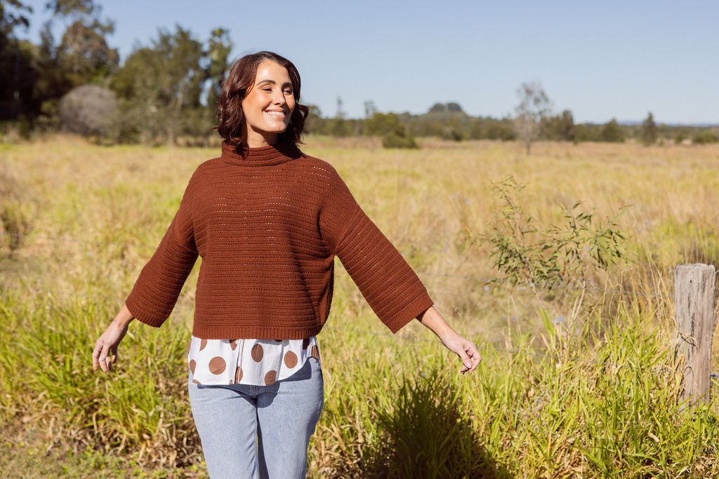 Woman wearing a See Saw copper pointelle sweater and polka dot shirt standing in a field with a clear blue sky.