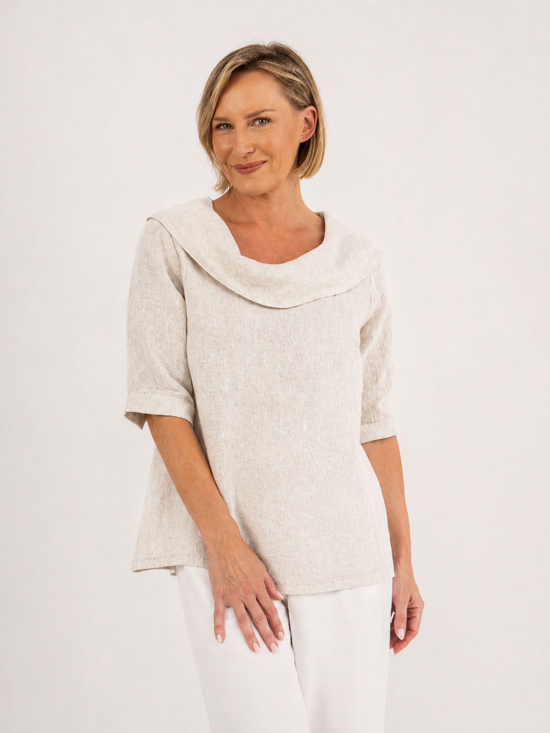 A woman modeling a natural linen See Saw top with a wide draped collar and elbow-length sleeves, paired with white pants, against a plain off-white background.