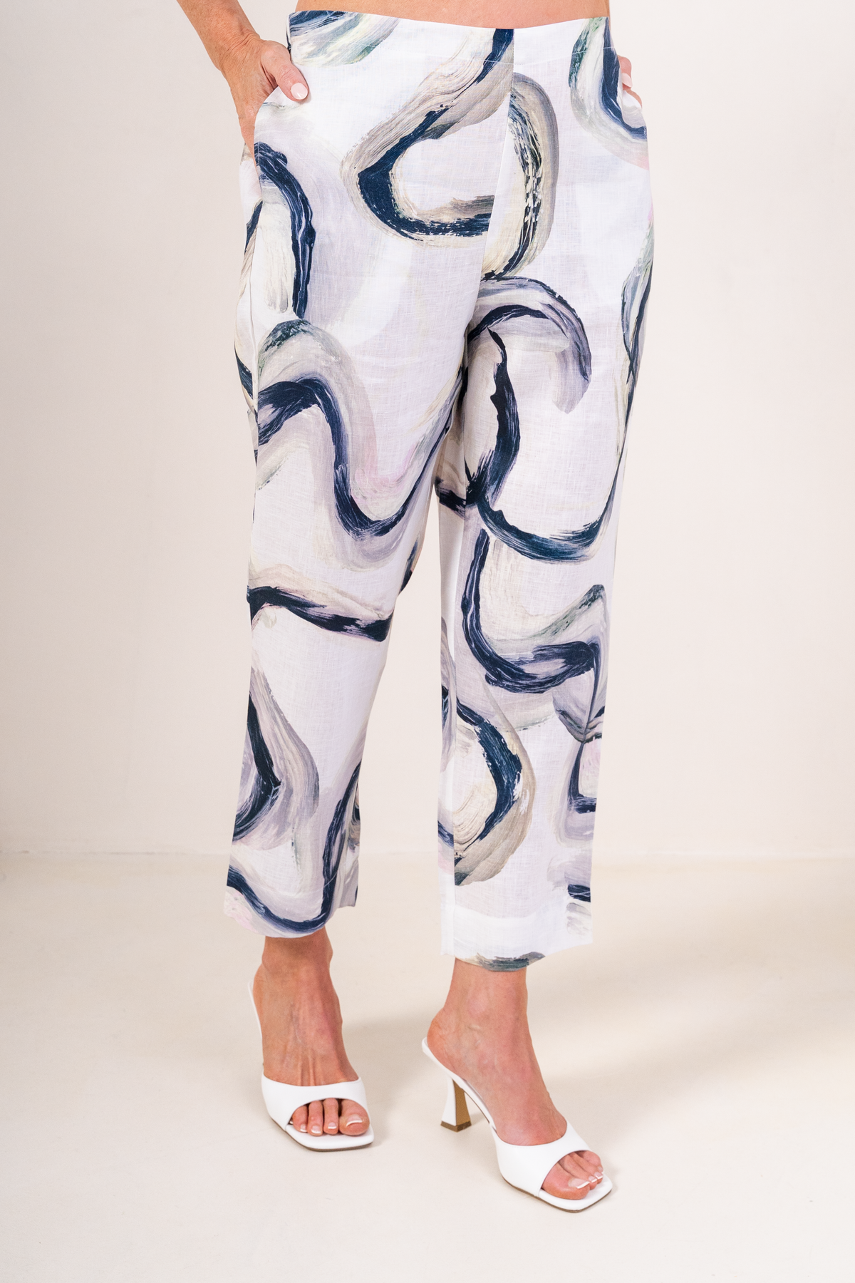 A model is shown from the waist down wearing white See Saw Flat front Riverstone Linen trousers, paired with white heeled sandals.