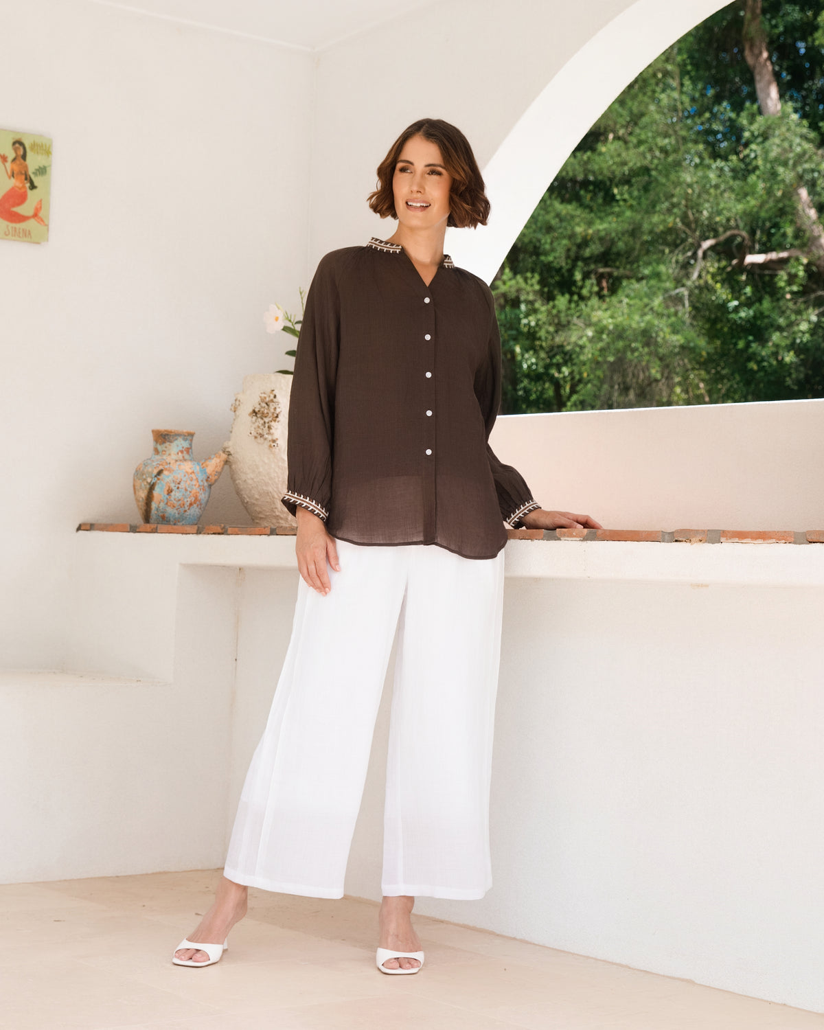 A woman modeling a See Saw Ramie brown button-front top and matching white wide-leg cropped pants, standing in a bright white architectural interior with an arched window showing greenery outside.