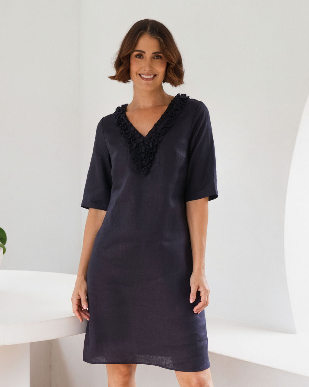 A woman modeling a short-sleeve See Saw linen dress in dark navy blue, featuring a decorative ruffled V-neckline, set against a bright, minimalist white background.