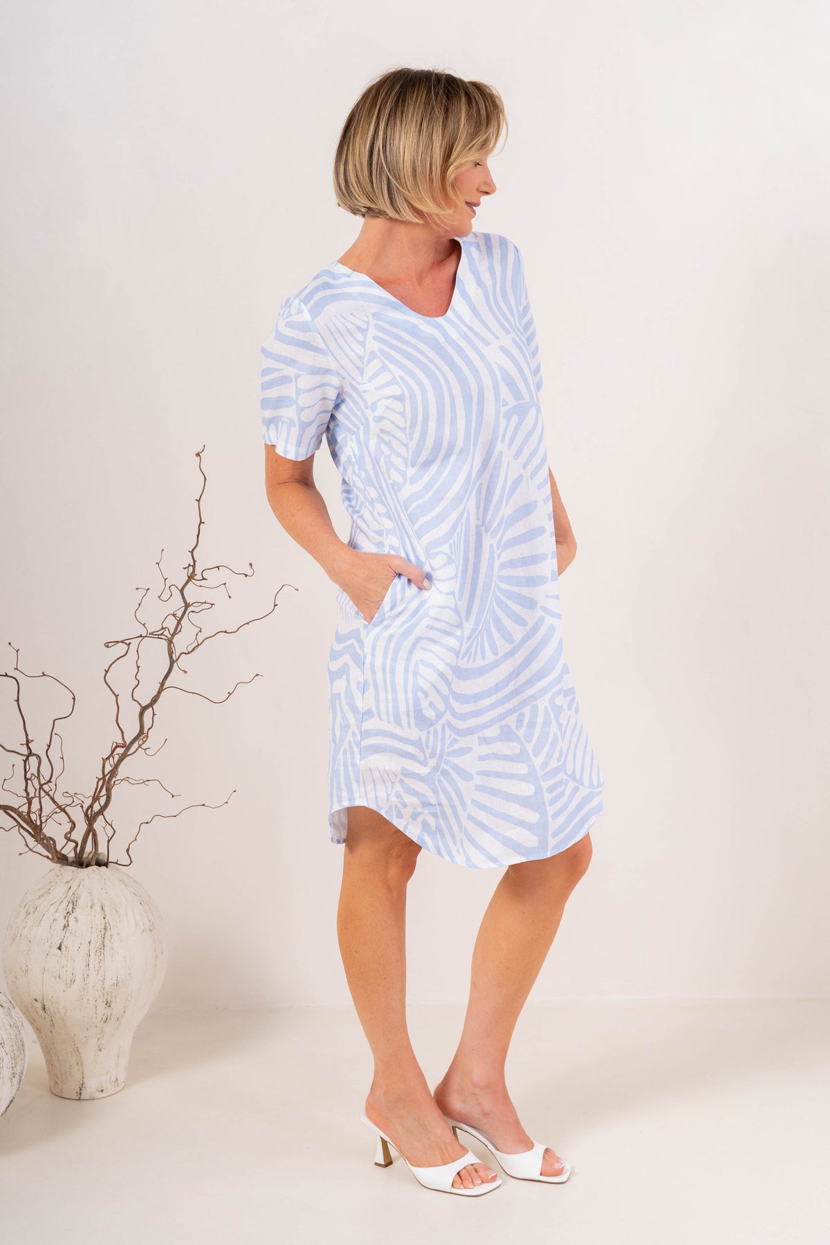 A woman modeling a light blue and white patterned short-sleeve dress with pockets, standing in a studio setting with a decorative vase on the left.