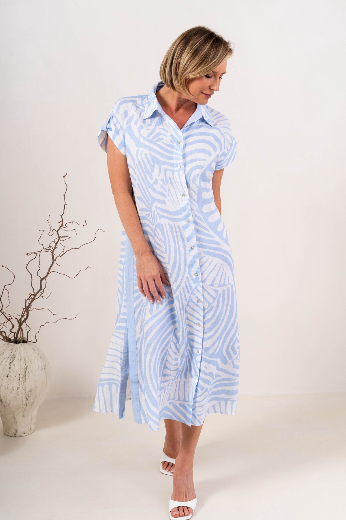 A woman modeling a Palm Breeze Linen See Saw abstract patterned midi shirt dress with short sleeves, a button-down collar, and a front slit, standing in a studio setting.
