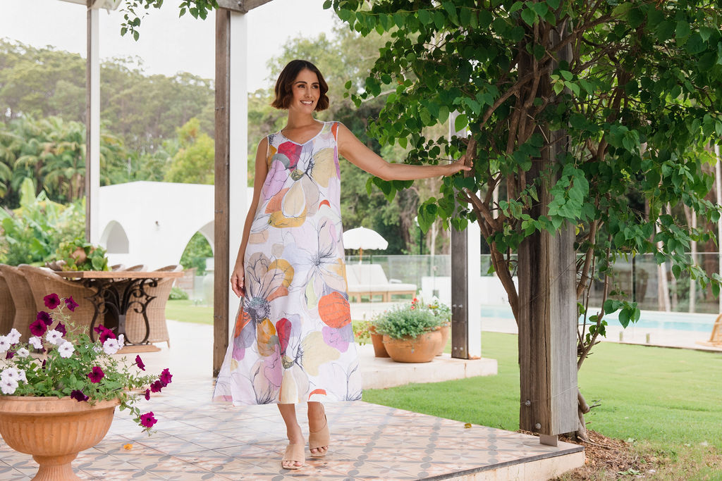 A woman wearing a sleeveless, linen See Saw knee-length A-line dress with a Petal Drift Print, standing on a tiled patio in a garden setting.