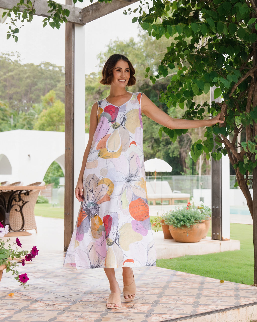 A woman wearing a sleeveless, linen See Saw knee-length A-line dress with a Petal Drift Print, standing on a tiled patio in a garden setting.