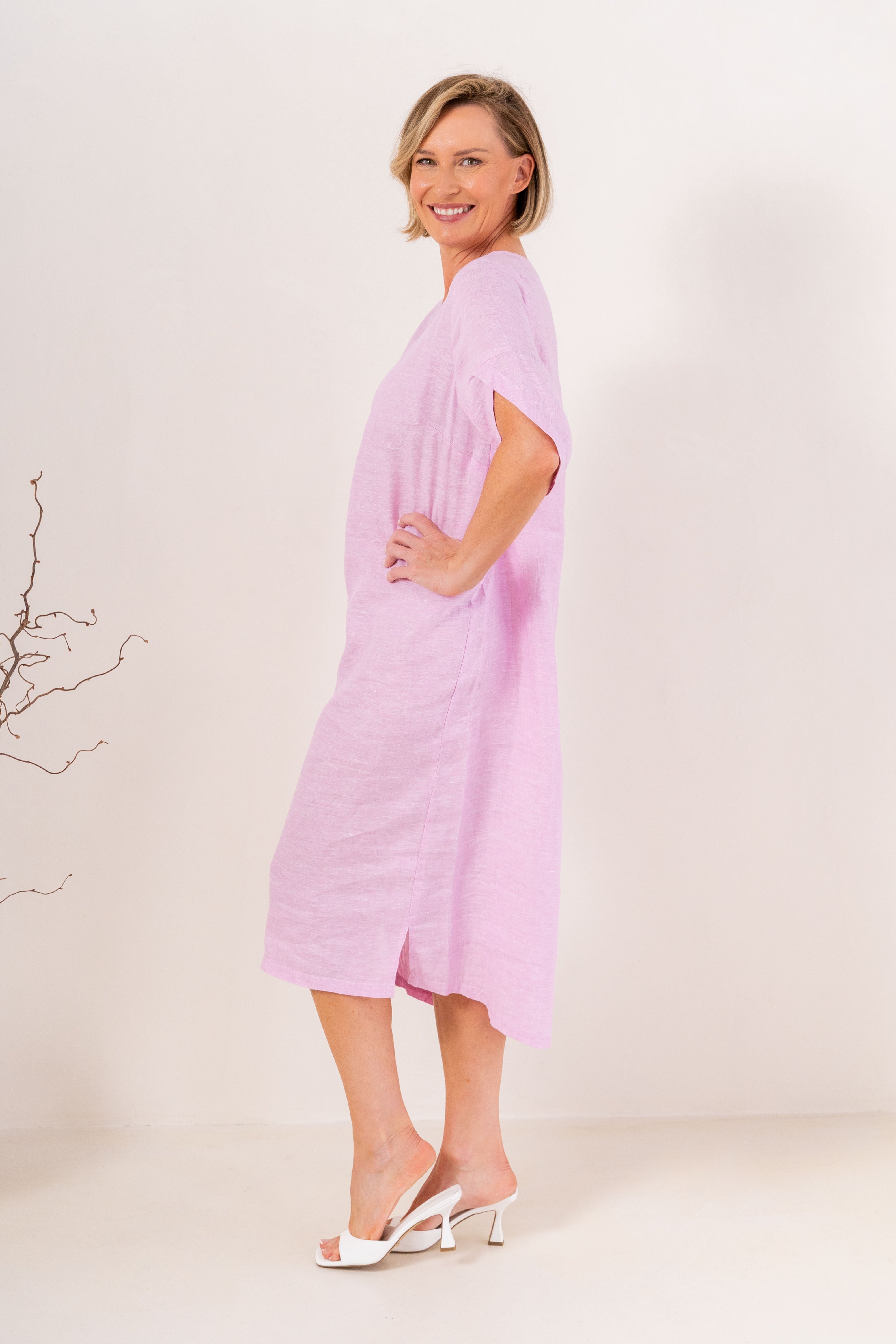 A woman modeling a See Saw Linen pink short-sleeve midi dress with a relaxed fit, standing against a plain off-white studio background.