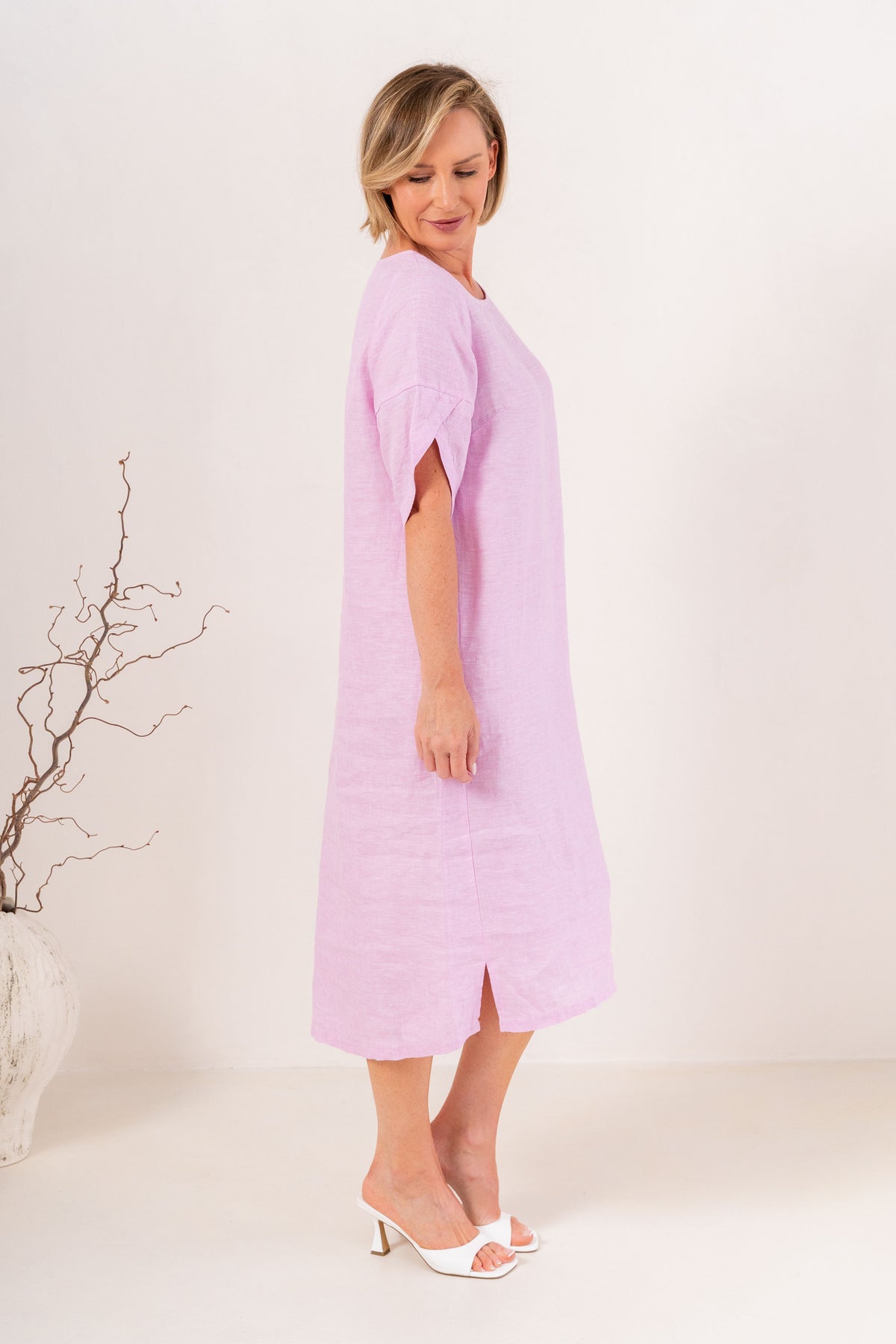 A woman modeling a See Saw Linen pink short-sleeve midi dress with a relaxed fit, standing against a plain off-white studio background.