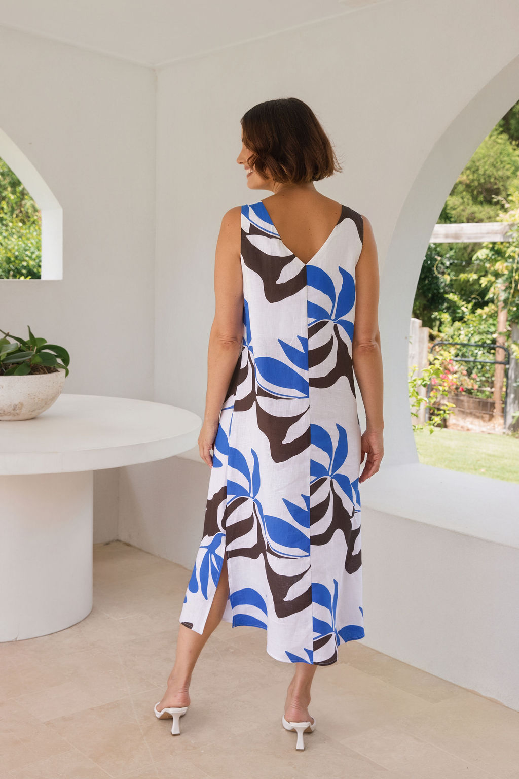 A woman models a sleeveless See Saw midi dress with a Shadow Leaf pattern in blue, brown, and white, standing in a bright, modern white architectural setting.