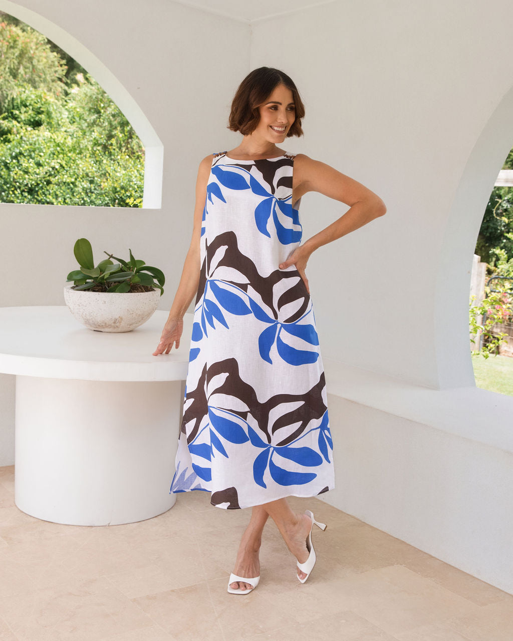 A woman models a sleeveless See Saw midi dress with a Shadow Leaf pattern in blue, brown, and white, standing in a bright, modern white architectural setting.