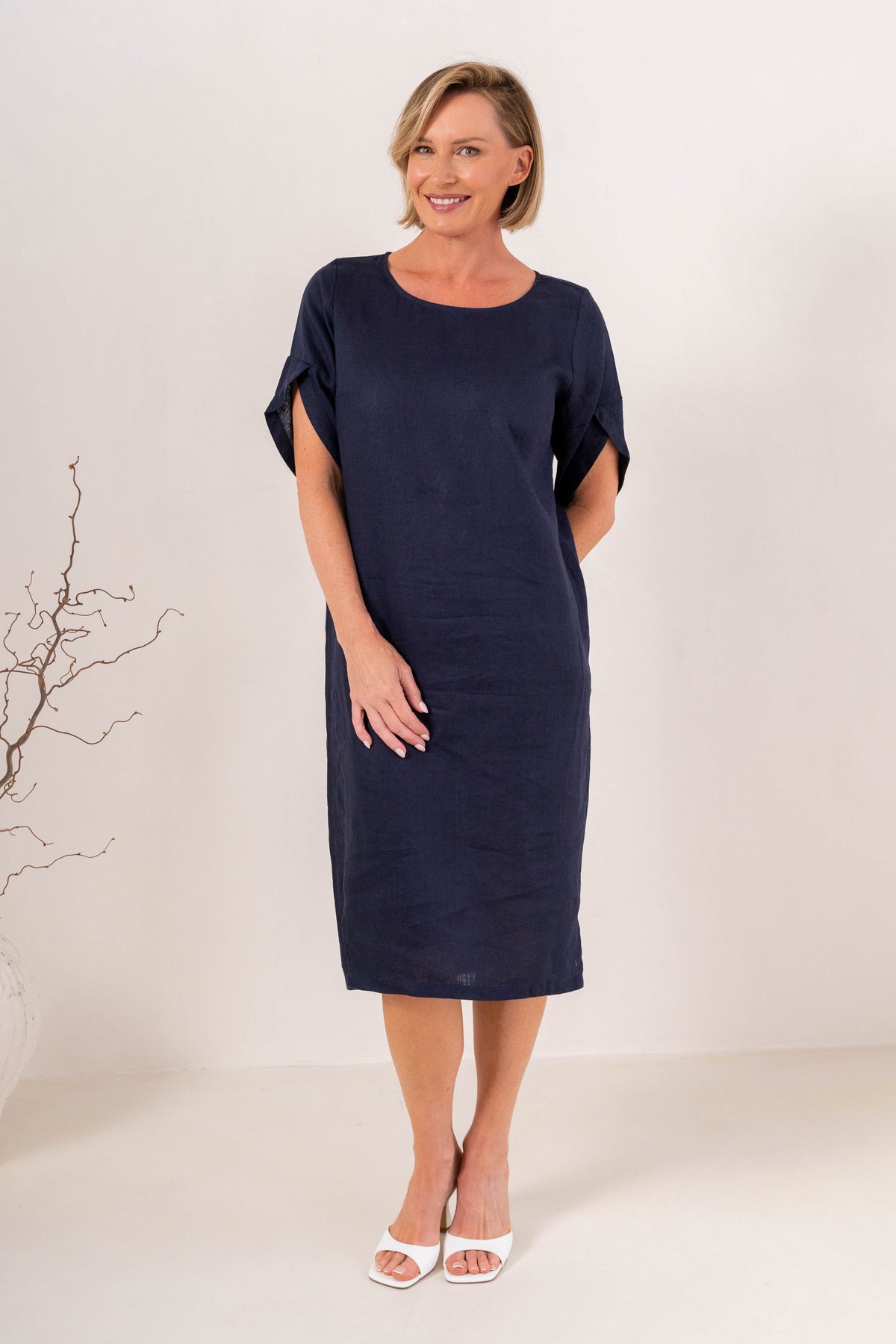 A woman modeling a See Saw Linen navy blue short-sleeve midi dress with a relaxed fit, standing against a plain off-white studio background.