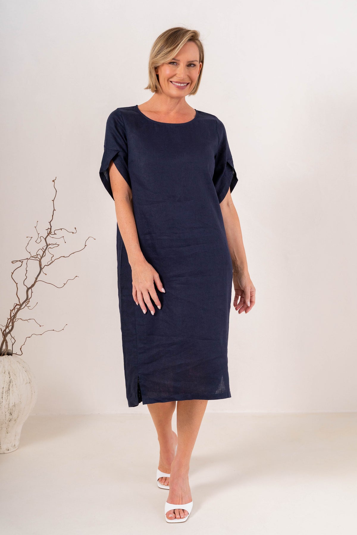 A woman modeling a See Saw Linen navy blue short-sleeve midi dress with a relaxed fit, standing against a plain off-white studio background.