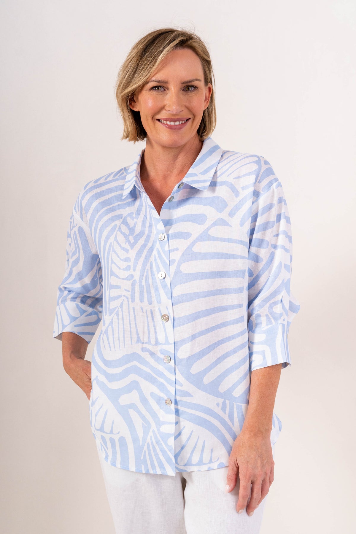A woman modeling a light blue and white linen See Saw patterned button-up shirt with three-quarter sleeves, paired with white pants, against a plain light background.