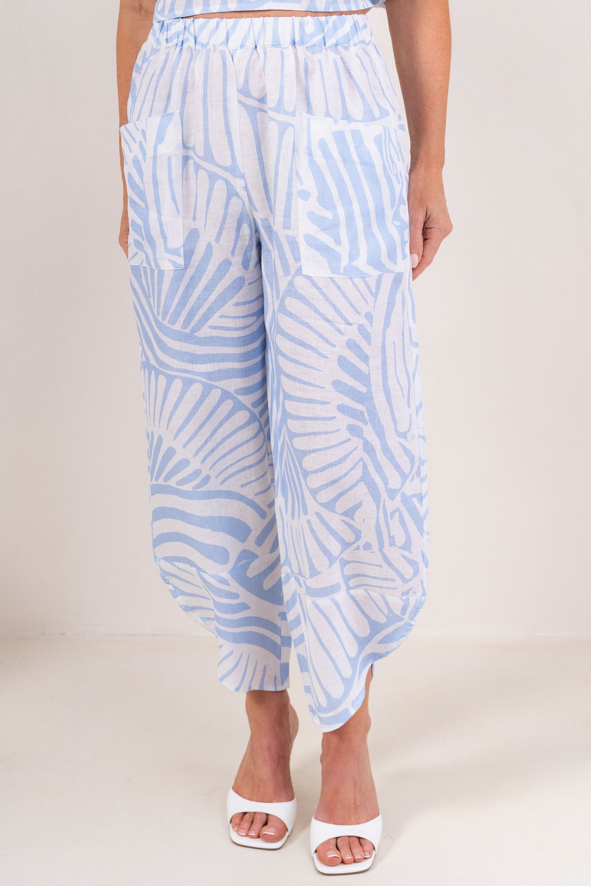 A person wearing loose, wide-leg cropped pants with a light blue and white Palm Breeze pattern linen See Saw brand, paired with white heeled sandals, shown against a plain light beige studio background.
