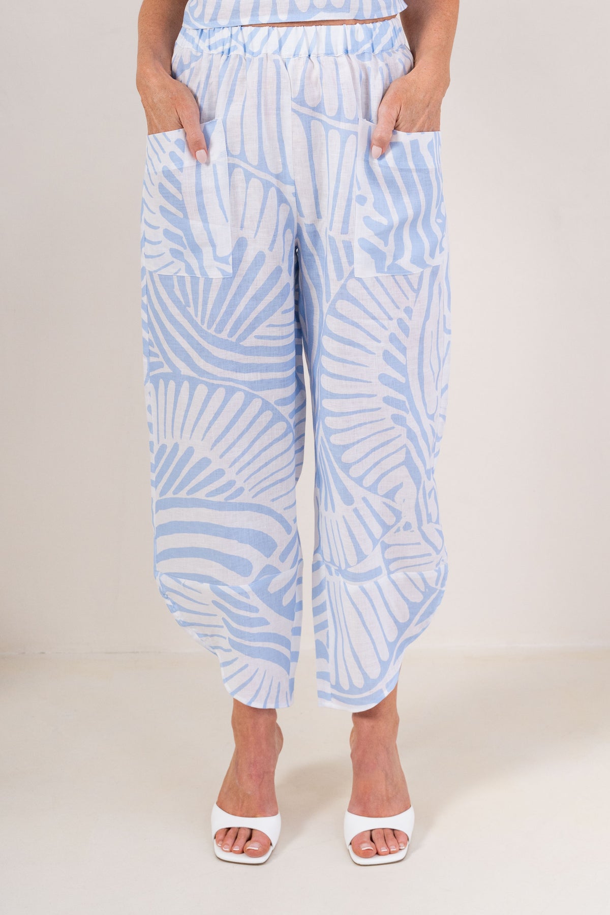 A person wearing light blue and white See Saw Palm Breeze print linen pants with side pockets, paired with white heeled sandals, shown against a plain light beige studio background.