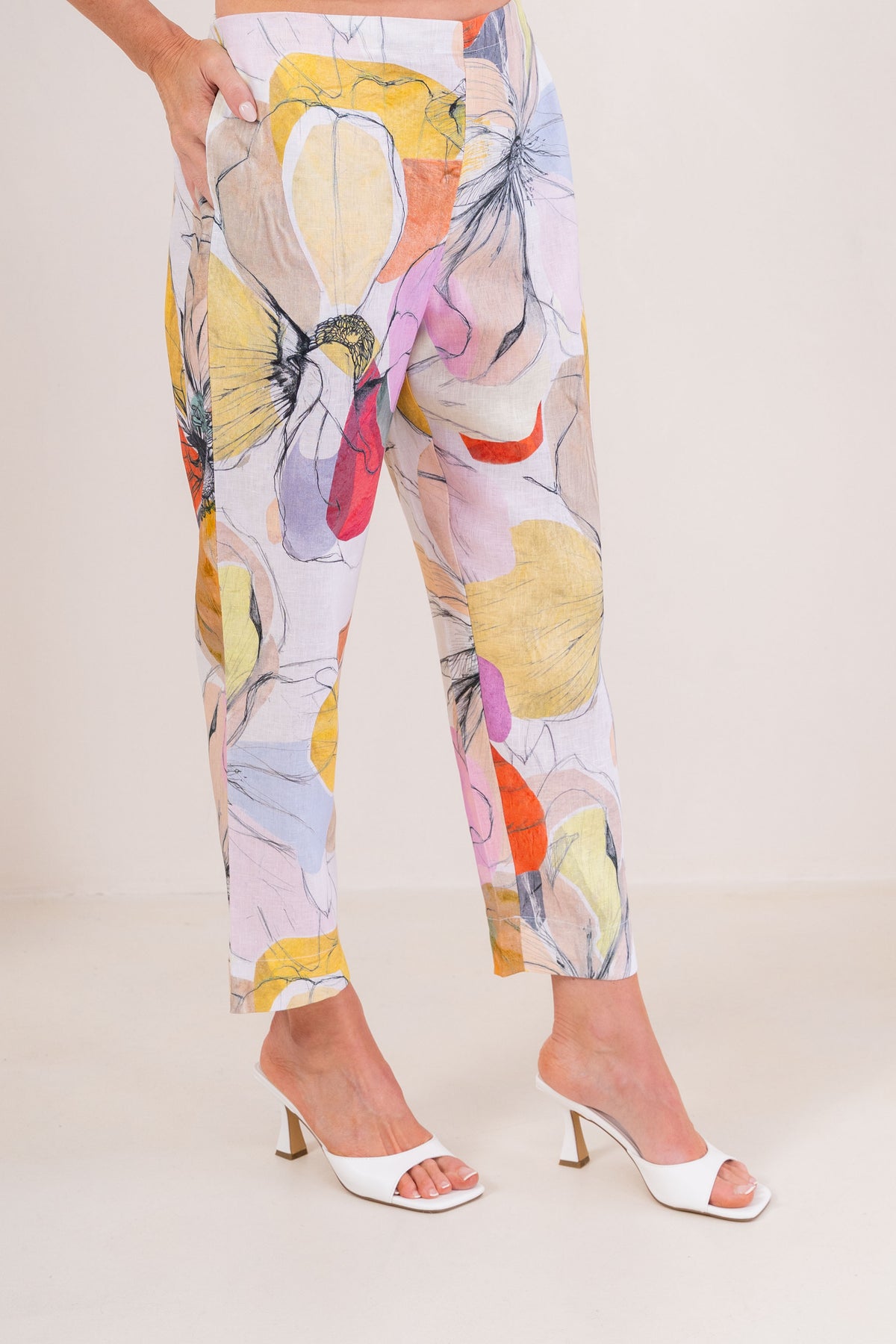 A person wearing See Saw linen cropped pants with a large abstract Petal Drift print in yellow, pink, orange, purple, and blue on a light base, paired with white heeled sandals against a plain beige studio background.
