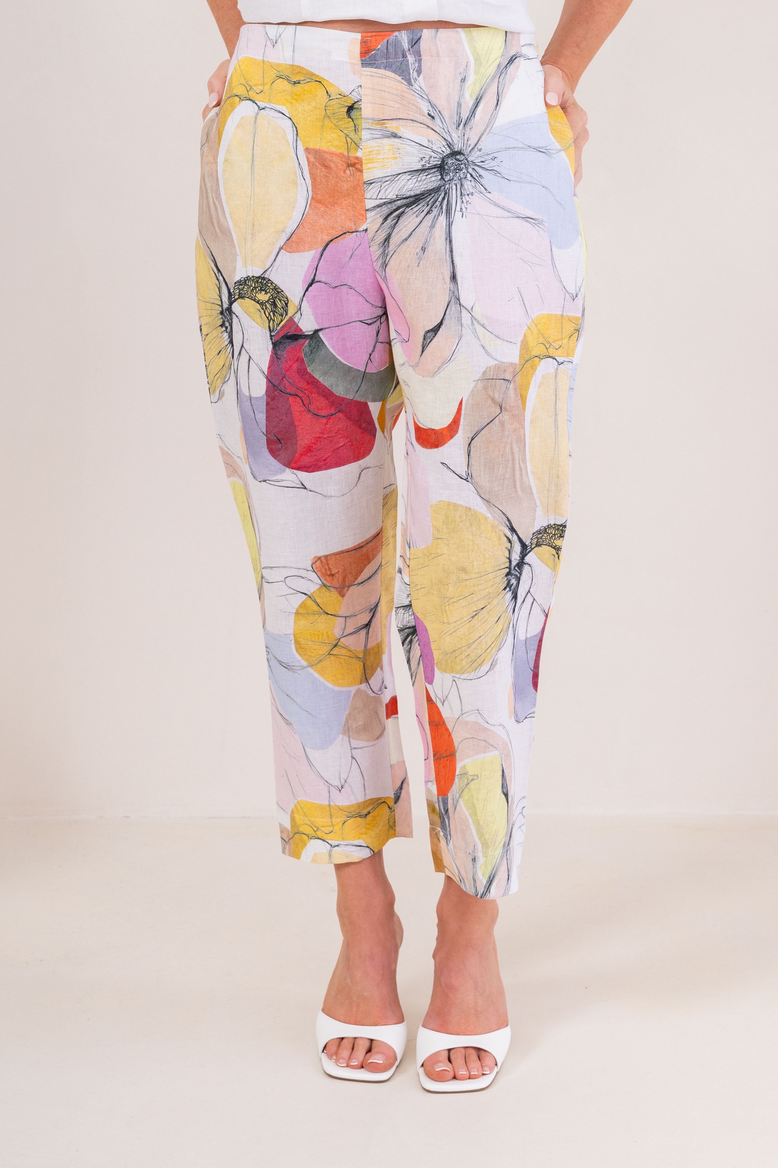 A person wearing See Saw linen cropped pants with a large abstract Petal Drift print in yellow, pink, orange, purple, and blue on a light base, paired with white heeled sandals against a plain beige studio background.