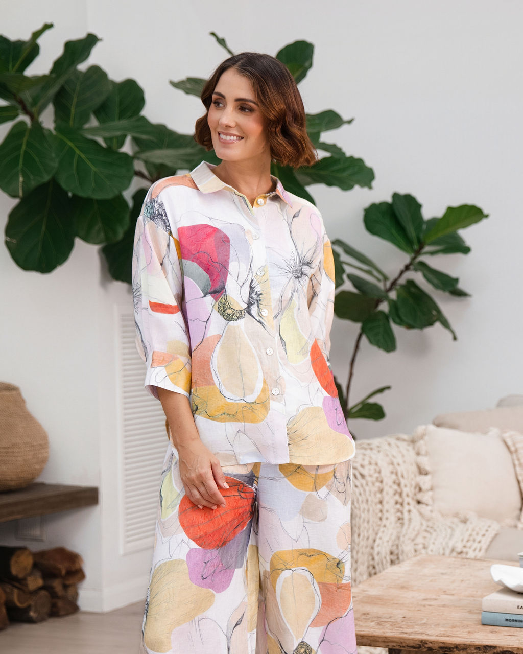 A woman modeling a matching two-piece See Saw linen set with petal drift  floral print, standing in a cozy, modern living room setting with indoor plants and a sofa.