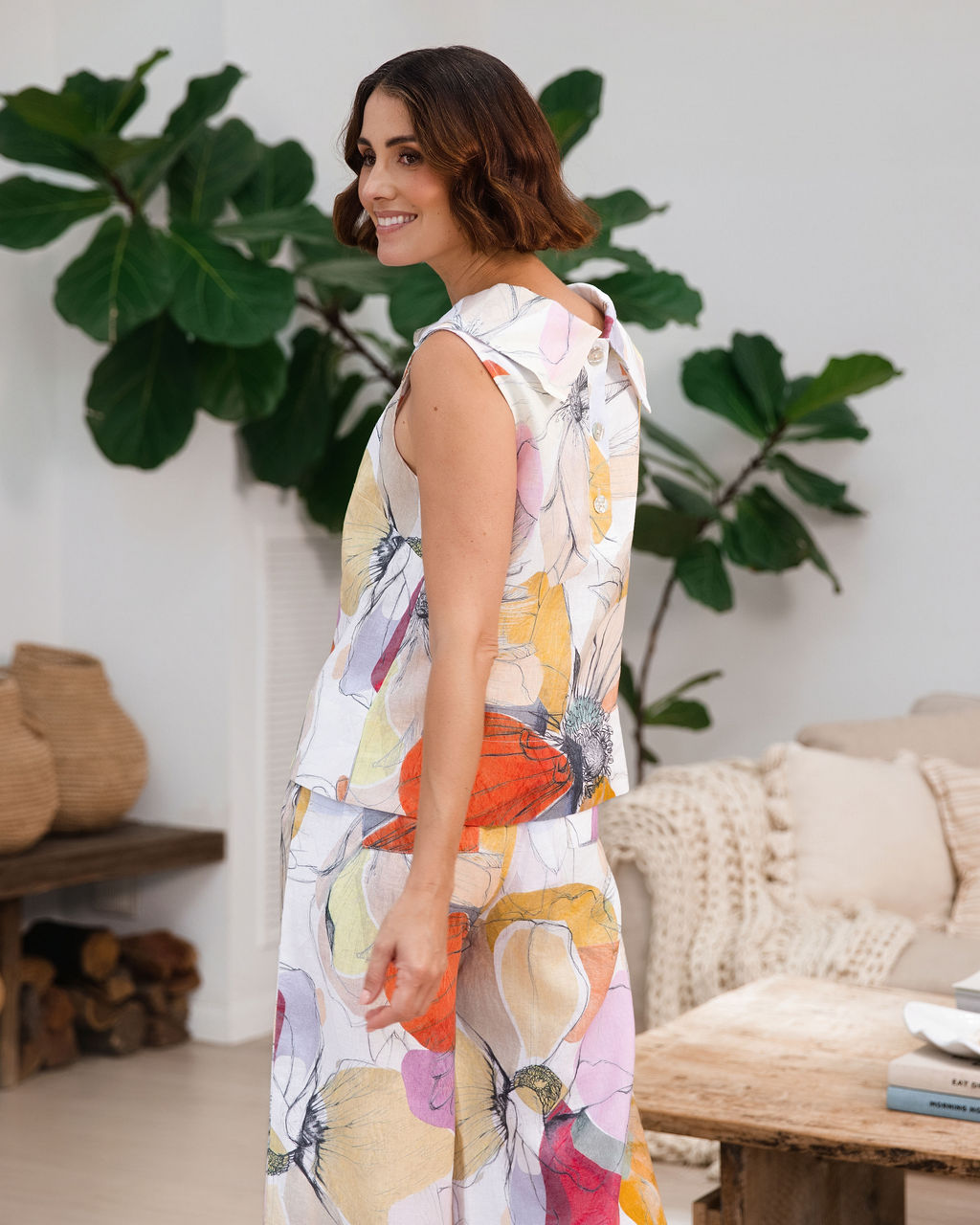 A woman stands in a bright, modern living room wearing a sleeveless white jumpsuit with a colorful See Saw linen abstract petal print in orange, pink, yellow, and blue. She is holding a small orange clutch.