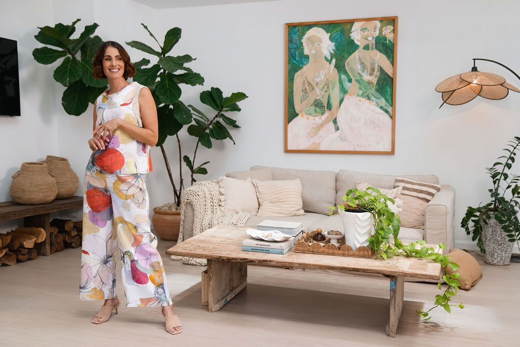 A woman stands in a bright, modern living room wearing a sleeveless white jumpsuit with a colorful See Saw linen abstract petal print in orange, pink, yellow, and blue. She is holding a small orange clutch.
